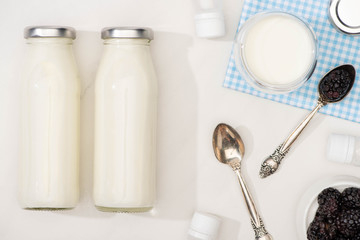 Top view of bottles and glass of yogurt on cloth, teaspoons, blackberries and containers with starter cultures on white