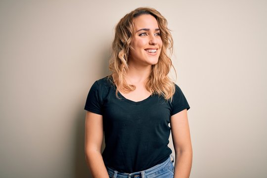 Young beautiful blonde woman wearing casual t-shirt standing over isolated white background looking away to side with smile on face, natural expression. Laughing confident.