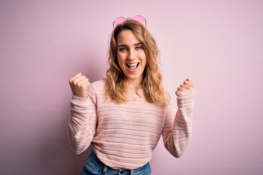 Young beautiful blonde woman wearing casual sweater and sunglasses over pink background celebrating surprised and amazed for success with arms raised and open eyes. Winner concept.