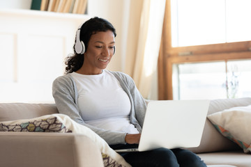 Fototapeta premium African american girl wearing headphones work at laptop while sit on sofa in the living room, online shopping on Internet, chatting with friends on social networks, typing email or message