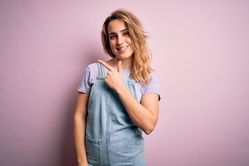 Young beautiful blonde woman wearing casual denim overalls standing over pink background cheerful with a smile of face pointing with hand and finger up to the side with happy and natural expression