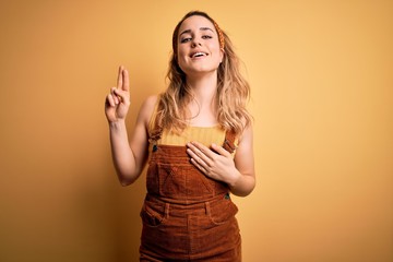 Young beautiful blonde woman wearing overalls and diadem standing over yellow background smiling...