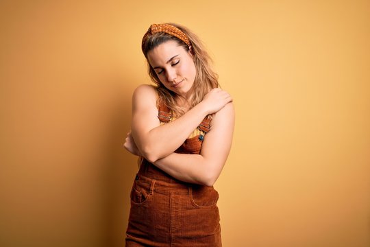 Young beautiful blonde woman wearing overalls and diadem standing over yellow background Hugging oneself happy and positive, smiling confident. Self love and self care