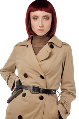 A young lady is wearing a natural look bricky-coloured wig with bangs. The lady with short blunt bob in a buttoned beige coat and a beige turtleneck is posing with a black gun in her leather belt.  