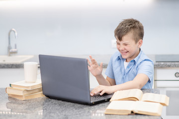A boy with a blue shirt waves his hand in greeting at the laptop camera. The student is engaged remotely on a laptop with a teacher. Distance Education Concept