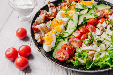 Cobb salad with chicken, avocado, tomatoes, eggs, bacon and cheese dressed with sauce on a white wooden  table close up.