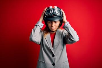 Young beautiful blonde motorcyclist woman wearing motorcycle helmet over red background suffering from headache desperate and stressed because pain and migraine. Hands on head.