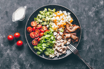 Cobb salad with chicken, avocado, tomatoes, eggs, bacon and cheese with sauce on a black  table top view