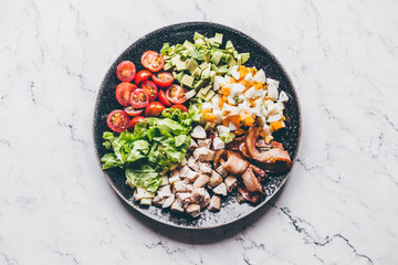 American traditional Cobb salad with chicken, avocado, tomatoes, eggs, bacon and cheese in a black plate on a marble table.