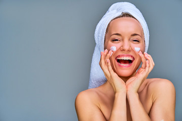Girl with smile laughing in towel applying a white cream on her cheeks by fingers. Beauty concept