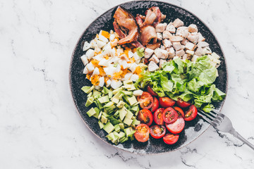 American traditional Cobb salad with chicken, avocado, tomatoes, eggs, bacon and cheese in a black plate on a marble table.