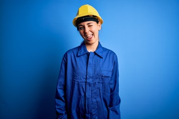 Young beautiful worker woman with blue eyes wearing security helmet and uniform winking looking at the camera with sexy expression, cheerful and happy face.