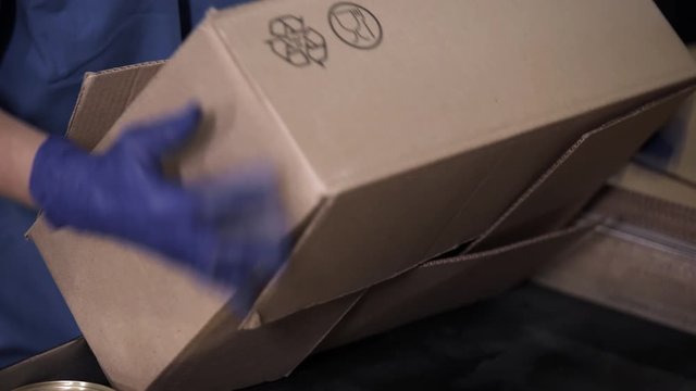 Female hands in blue protective gloves packs cans of meat in boxes at factory. Meat cans being sealed and tightened in food processing plant.