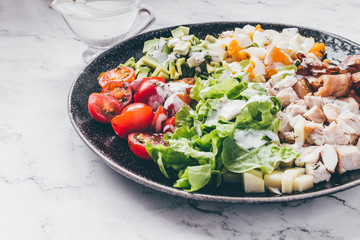 Cobb salad with chicken, avocado, tomatoes, eggs, bacon and cheese dressed with sauce on a marble table close up.
