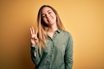 Young beautiful blonde woman with blue eyes wearing green shirt over yellow background showing and pointing up with fingers number three while smiling confident and happy.