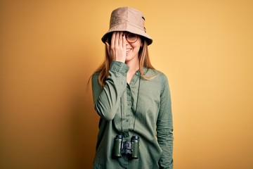 Beautiful blonde explorer woman with blue eyes wearing hat and glasses using binoculars covering one eye with hand, confident smile on face and surprise emotion.