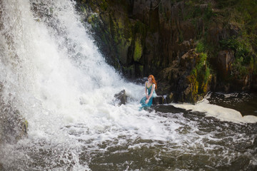 beautiful red-haired girl bathes in a stormy stream of a waterfall, hot summer