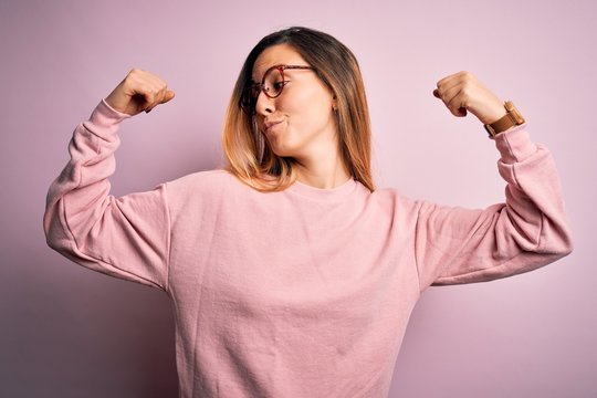 Beautiful blonde woman with blue eyes wearing sweater and glasses over pink background showing arms muscles smiling proud. Fitness concept.