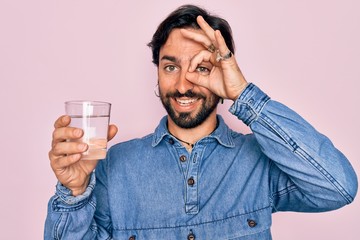 Young handsome hispanic bohemian hipster man with bear drinking a fresh glass of water with happy face smiling doing ok sign with hand on eye looking through fingers