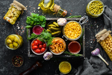 Pickled vegetables and mushrooms in glass jars in Wooden box on black stone background. Top view.