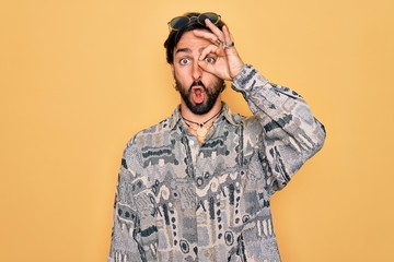 Young handsome hispanic bohemian man wearing hippie style and sunglasses doing ok gesture shocked with surprised face, eye looking through fingers. Unbelieving expression.