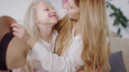 Slowmo of happy mother sitting on couch and hugging adorable little girl. She is taking off wide brim hat from her head and kissing her forehead, then they are looking at camera and smiling