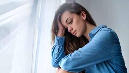 Young sad lonely pensive woman in a shirt holds his head with hand sits alone at home by the window during the depression and worries