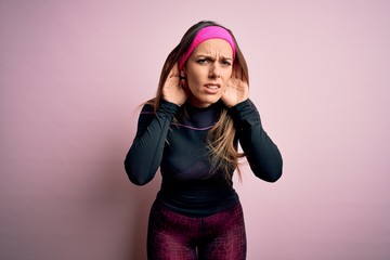 Young blonde fitness woman wearing sport workout clothes over isolated background Trying to hear both hands on ear gesture, curious for gossip. Hearing problem, deaf