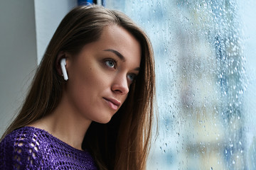 Young pensive woman music lover with wireless earbuds listens to soothing calming relaxing music during standing by the window with raindrops in rainy autumn weather