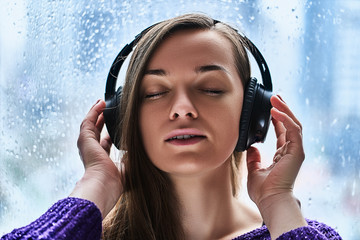 Woman music lover with closed eyes in wireless headphones enjoys and listens to soothing calming relaxing music during rainy fall weather