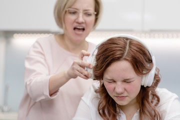 Mom scolds the teenager's daughter while she sits with headphones and ignores the mother. Mom tries to remove the headphones from her daughter to shout to her, the daughter closed her eyes in fear.