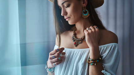 Stylish brunette boho chic woman wears white blouse and straw hat with big earrings, bracelets,...