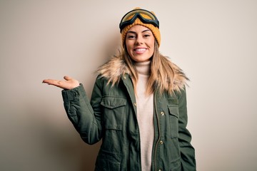 Young brunette skier woman wearing snow clothes and ski goggles over white background smiling cheerful presenting and pointing with palm of hand looking at the camera.