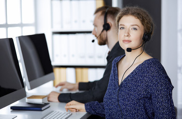 Business woman talking by headset while sitting with red-bearded colleague in modern office....