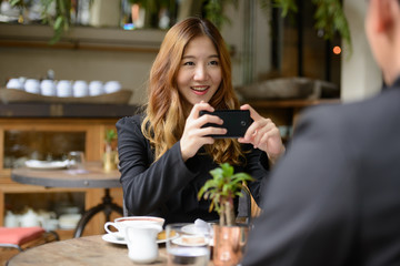 Happy young Asian business couple together at the coffee shop with woman taking picture using phone