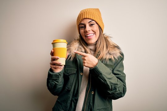 Young beautiful brunette woman wearing snow clothes drinking cup of coffee very happy pointing with hand and finger