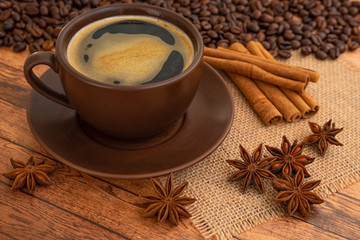 Coffee with foam in a dark brown Cup and saucer on a napkin on a wooden table with scattered coffee beans, star anise and cinnamon.