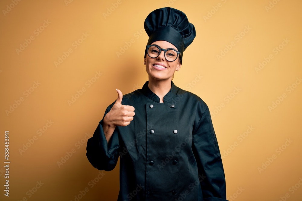 Sticker young beautiful brunette chef woman wearing cooker uniform and hat over yellow background doing happ