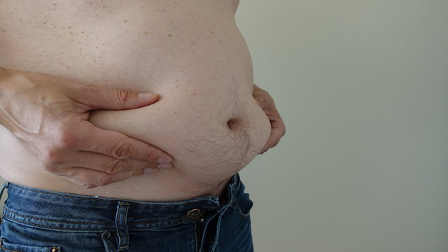 Overweight Man Squishing Belly Fat With Both Hands Wearing Blue Jeans Standing On Left Side Against Beige Background