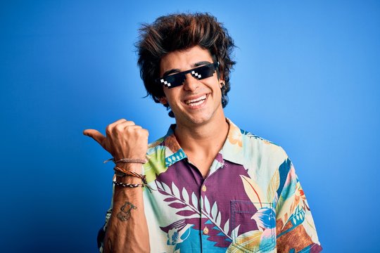 Young handsome man wearing thug life glasses standing over isolated blue background smiling with happy face looking and pointing to the side with thumb up.