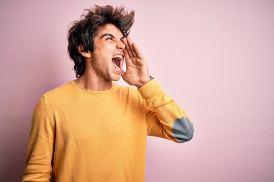 Young handsome man wearing yellow casual t-shirt standing over isolated pink background shouting and screaming loud to side with hand on mouth. Communication concept.