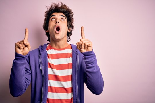 Young man wearing striped t-shirt and purple sweashirt over isolated pink background amazed and surprised looking up and pointing with fingers and raised arms.