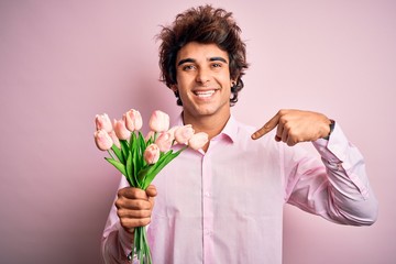 Young handsome man holding flowers standing over isolated pink background with surprise face pointing finger to himself