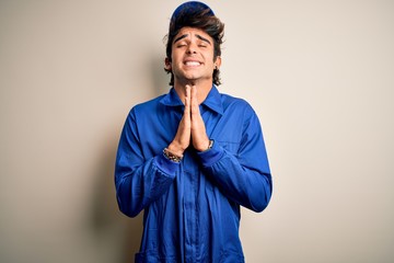 Young mechanic man wearing blue cap and uniform standing over isolated white background begging and praying with hands together with hope expression on face very emotional and worried. Begging.