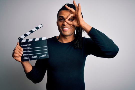 Young african american afro director man making movie using clapboard with happy face smiling doing ok sign with hand on eye looking through fingers