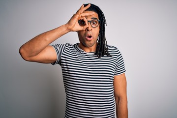 Young handsome african american man with dreadlocks wearing striped t-shirt and glasses doing ok gesture shocked with surprised face, eye looking through fingers. Unbelieving expression.