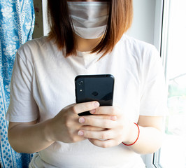 Medical mask on the face of an unknown girl, hands and mobile phone.The house near the window.