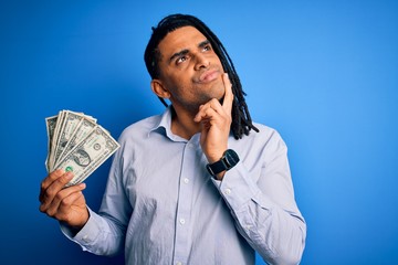 Young african american afro man with dreadlocks holding dollars over blue background serious face...