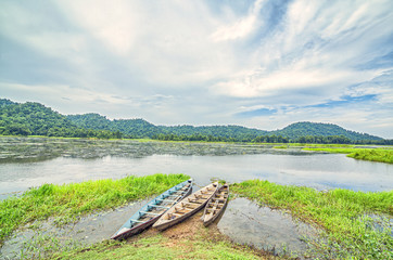 Three boats anchored to the banks of a Lake with hills visible in thhe distance and a blue sky with white clouds, on a sunny day.