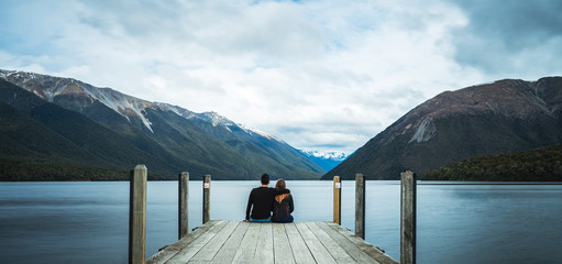 Rotoiti Lake, New Zealand, October 8, 2019: Beautiful picture of a loving couple holding each other admiring the lake and sitting at the end of a yetty with snowy mountains in the background